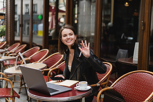 happy woman in black jacket holding smartphone and waving hand near laptop and cup of coffee in french outdoor cafe