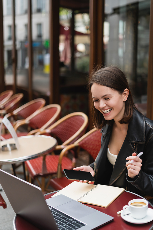 cheerful freelancer in black jacket holding smartphone and pen near laptop in french outdoor cafe