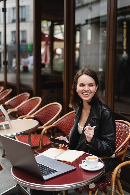 joyful in black jacket holding smartphone and pen near laptop and cup of coffee in french outdoor cafe