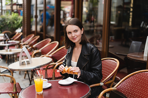 cheerful woman holding croissant near cup of coffee and glass of orange juice in outdoor cafe in paris