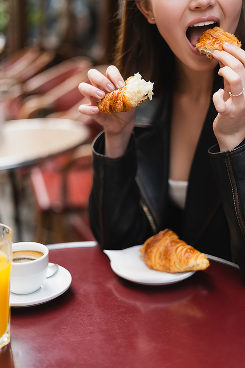 cropped view of woman eating croissant near cup of coffee and glass of orange juice in outdoor cafe in paris