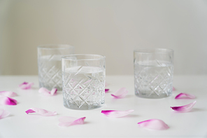 floral petals and faceted glasses with pure water on white tabletop isolated on grey