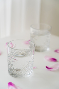 close up view of faceted glass with clean water near floral petals on blurred grey background