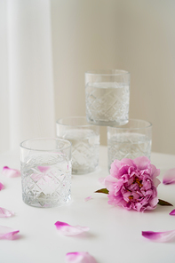 faceted glasses stacked near pink peony and petals on white tabletop and grey background