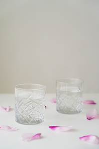 transparent glasses with fresh water near floral petals on white tabletop isolated on grey