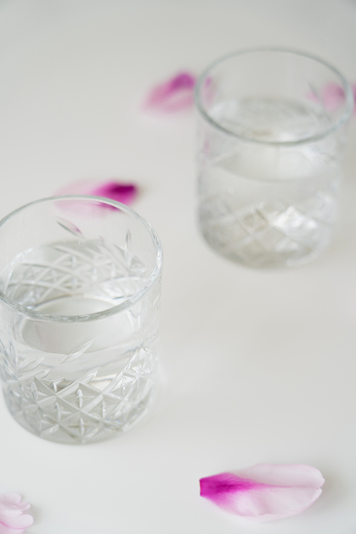close up view of glasses with fresh water near floral petals on white surface and blurred background