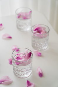 high angle view of faceted glasses with tonic and natural petals on white surface