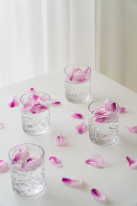 faceted glasses with gin tonic near floral petals scattered on white tabletop on grey background