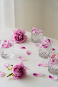 crystal glasses with tonic and floral petals near pink peonies on white tabletop and grey background