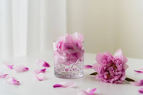 faceted glass with tonic and petals near pink peony on white tabletop and grey background