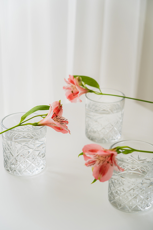 crystal glasses with water near alstroemeria flowers on white tabletop and grey background
