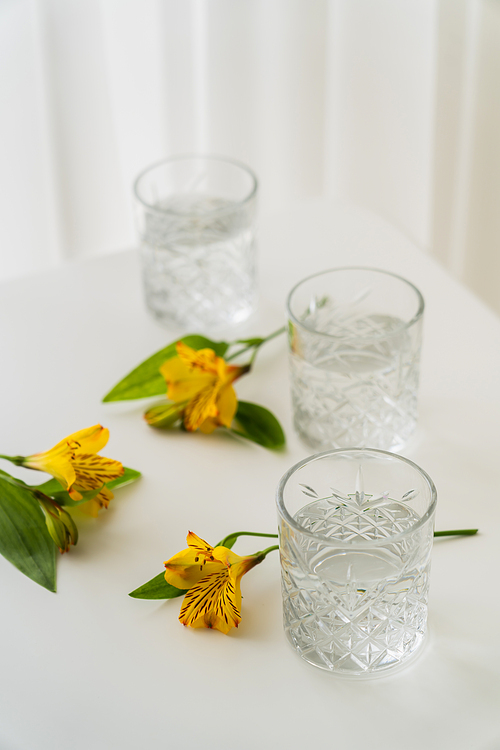 crystal glasses with water near yellow alstroemeria flowers on white tabletop and blurred background