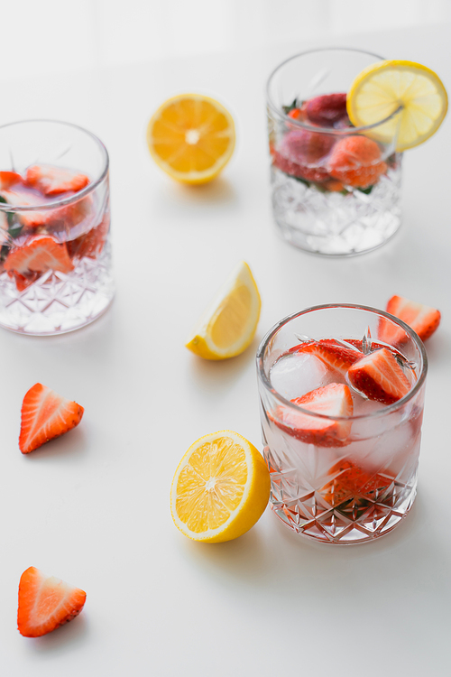 faceted glasses with fruit tonic drink near chopped strawberries and lemons on white tabletop