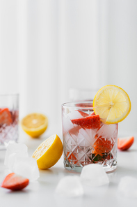 selective focus of tonic water with strawberries and ice near cut lemons on white blurred background