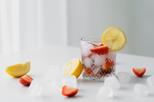 glass with cold strawberry tonic near chopped lemons and ice cubes on white surface and grey background