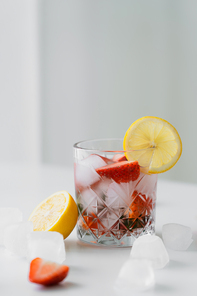 glass of iced tonic drink with chopped strawberries near cut lemon on white surface and grey background