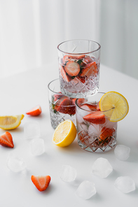 glasses with cold strawberry tonic near cut lemons and ice cubes on white tabletop