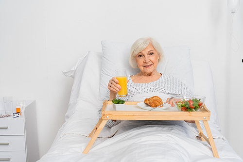 Senior patient holding orange juice near food on tray in hospital ward
