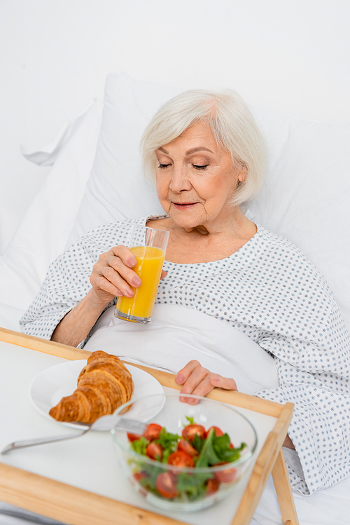 Elderly woman holding glass of orange juice near food on tray on hospital bed