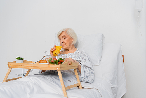 Elderly woman drinking orange juice near food on bed in hospital