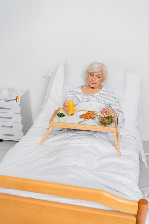 Elderly patient holding glass of orange juice near food in hospital ward