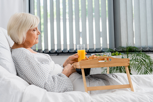 Side view of senior patient holding glass of orange juice near food in hospital