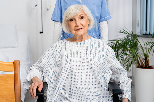Senior woman in wheelchair looking at camera near nurse in clinic