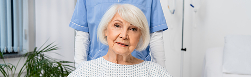 Senior patient looking at camera near blurred nurse in hospital, banner