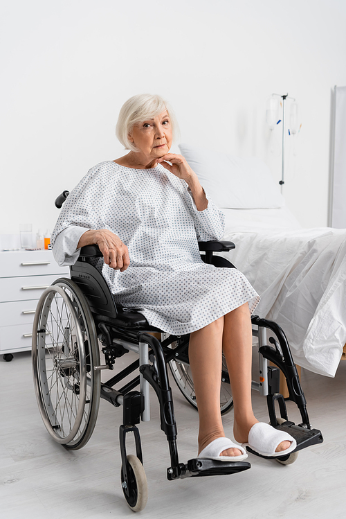 Elderly patient in gown looking at camera in wheelchair in clinic