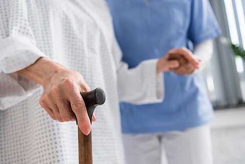 Cropped view of senior patient holding walking cane near blurred nurse