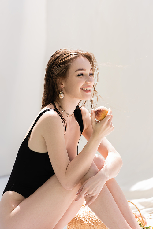 Positive young woman in swimsuit holding peach near handbag at resort