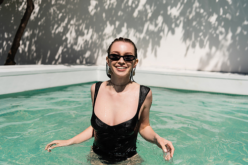 happy young woman with wet hair and stylish sunglasses sunbathing while swimming in pool