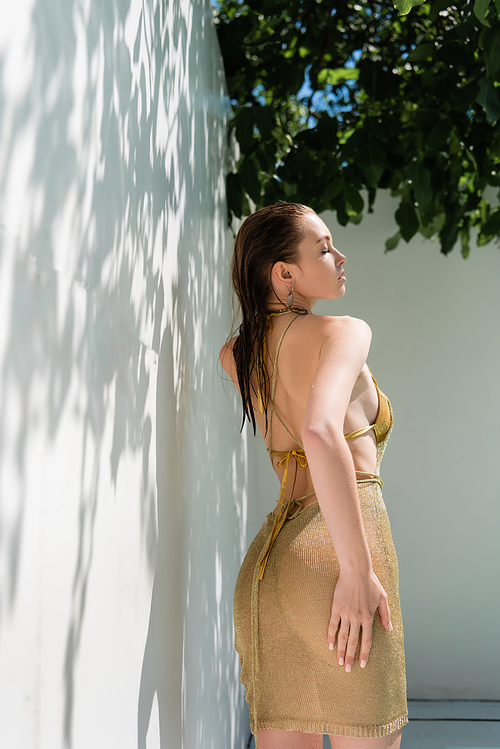 seductive woman in golden summer dress standing near while wall with shadows from green leaves