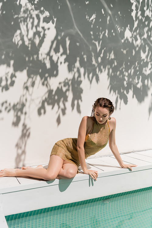 stylish woman in summer dress sitting near swimming pool and wall with shadows