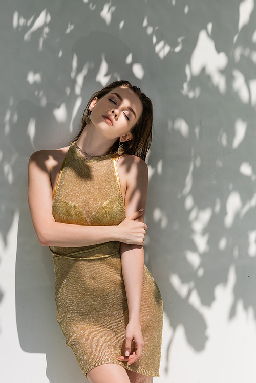 pretty young woman with closed eyes posing in summer dress near white wall with shadows from leaves