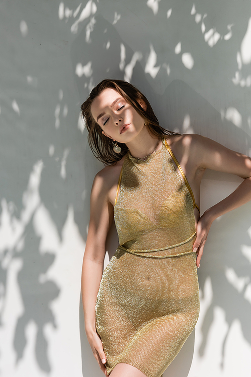 pretty young woman with closed eyes posing with hand on hip near white wall with shadows from leaves