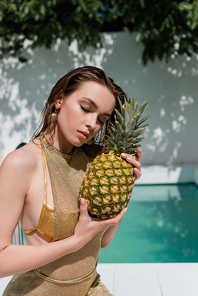 young woman with closed eyes in summer dress sitting with pineapple at poolside