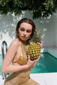 young woman in golden summer dress sitting with pineapple at poolside