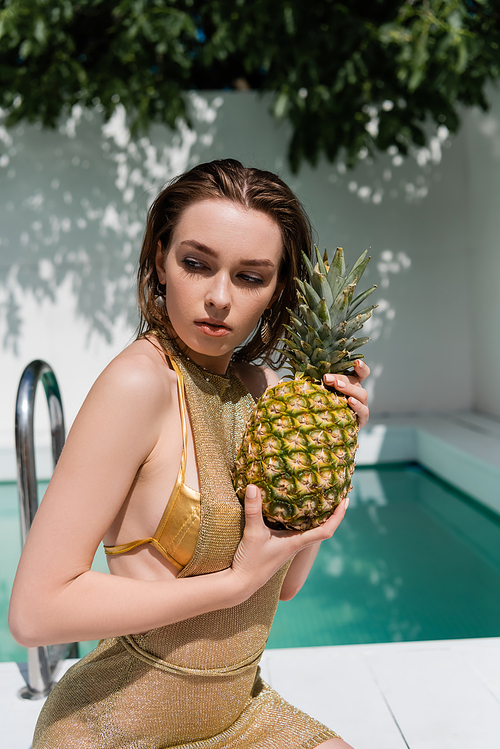 young woman in golden summer dress sitting with pineapple at poolside