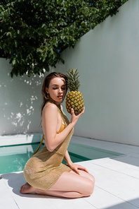 pretty young woman in golden summer dress sitting with fresh pineapple at poolside