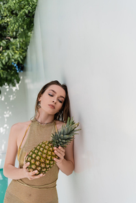 pretty young woman in golden summer dress holding ripe pineapple near white wall