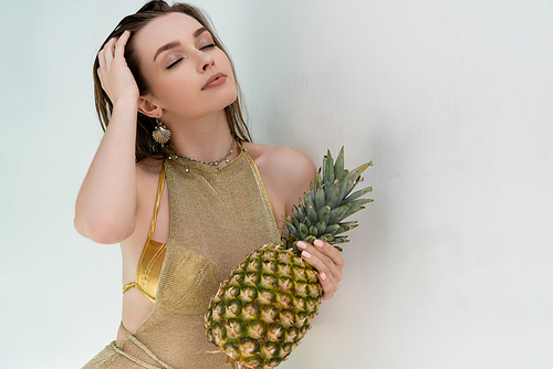 young woman in golden summer dress holding ripe pineapple near white wall