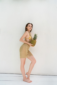 full length of young woman in golden summer dress holding fresh pineapple near white wall