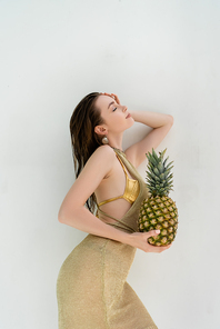 side view of smiling woman in golden swimwear and dress holding ripe pineapple near while wall