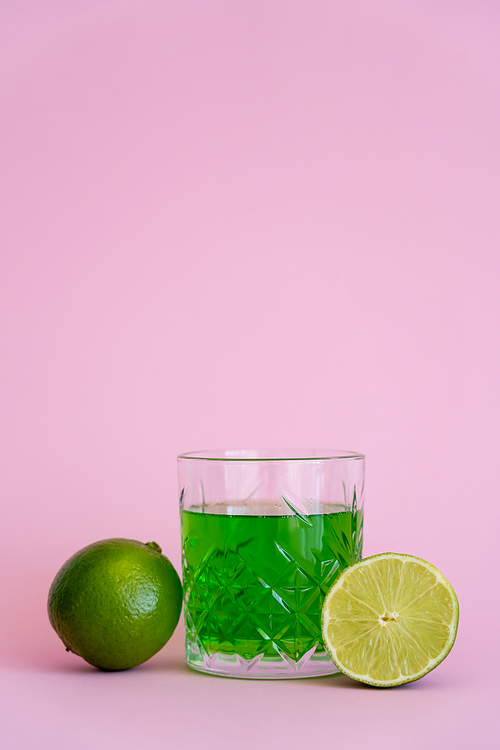 green alcohol drink in faceted glass near fresh limes on pink background