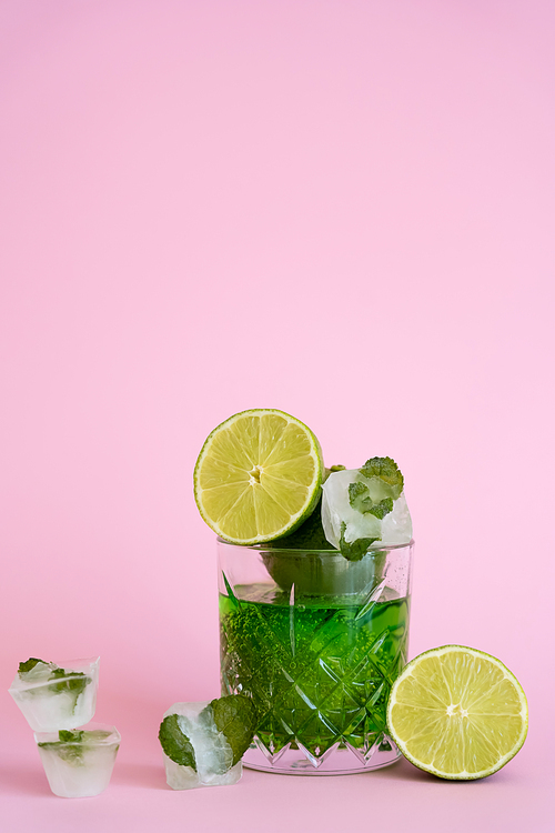 green and fresh limes in faceted glass with sparkling alcohol drink near ice cubes with peppermint on pink