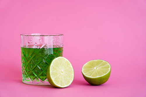 green alcohol drink in faceted glass with water drops near limes on pink
