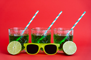 green alcohol drink in faceted glasses with straws and halves of limes near sunglasses on red