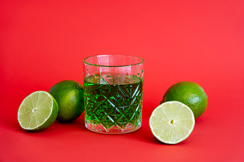 green alcohol drink in faceted glass near sour limes on red