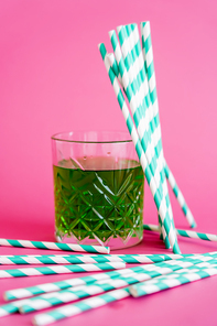 faceted glass of green alcohol drink near striped paper straws on pink
